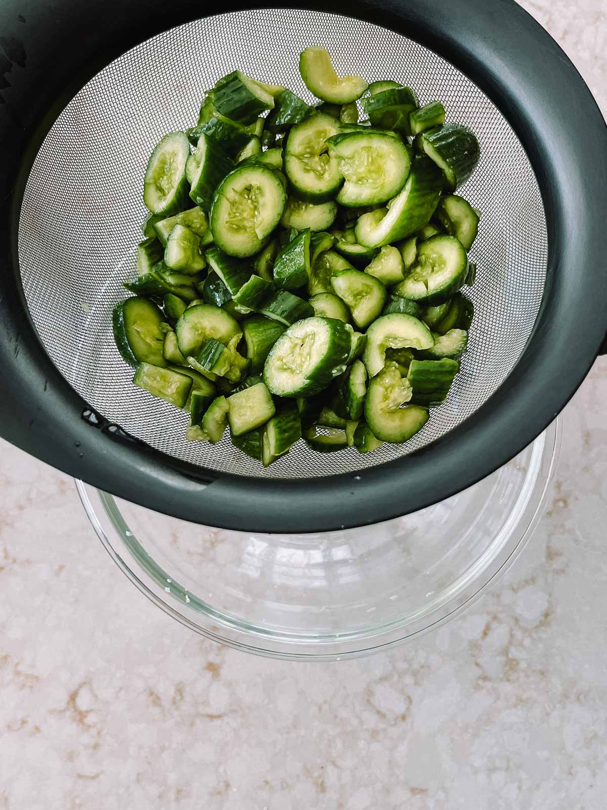 Sliced cucumbers in a strainer over a glass bowl.