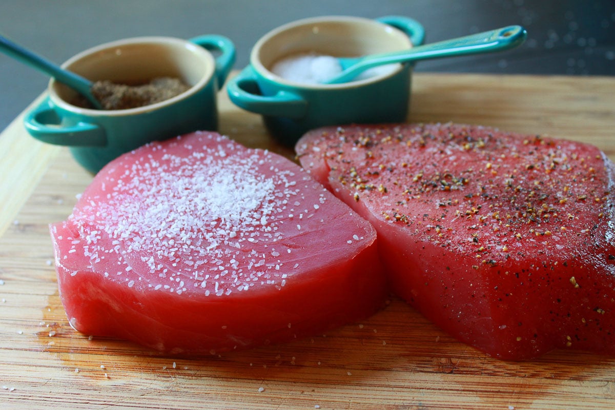 Two raw ahi tuna steaks on a wooden cutting board with small blue jars of salt and pepper in the background.