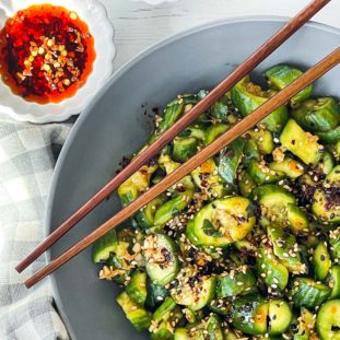 Smashed cucumber salad in a gray bowl with wooden chopsticks on top and a small white bowl of chili oil and a checkered napkin on the side.