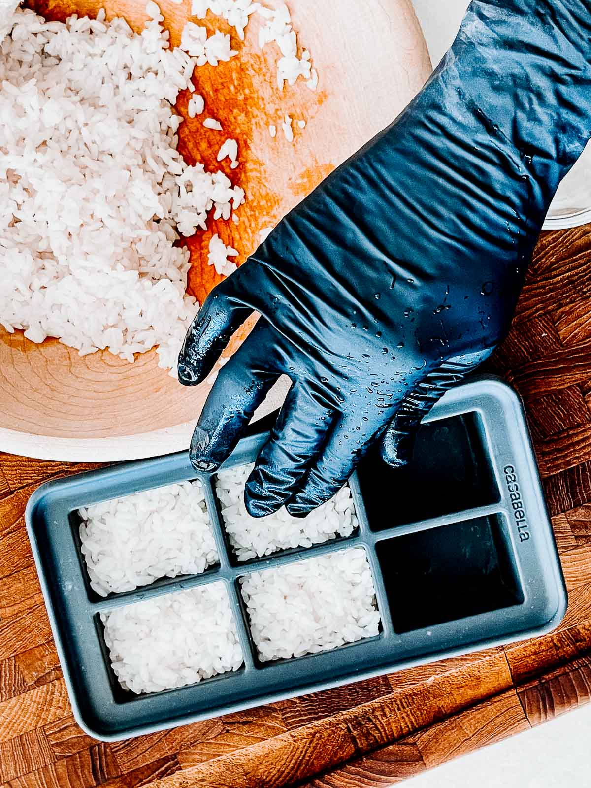 A hand wearing a black glove pressing sushi rice into a black silicone ice cube tray with a wooden bowl filled with sushi rice on the side.