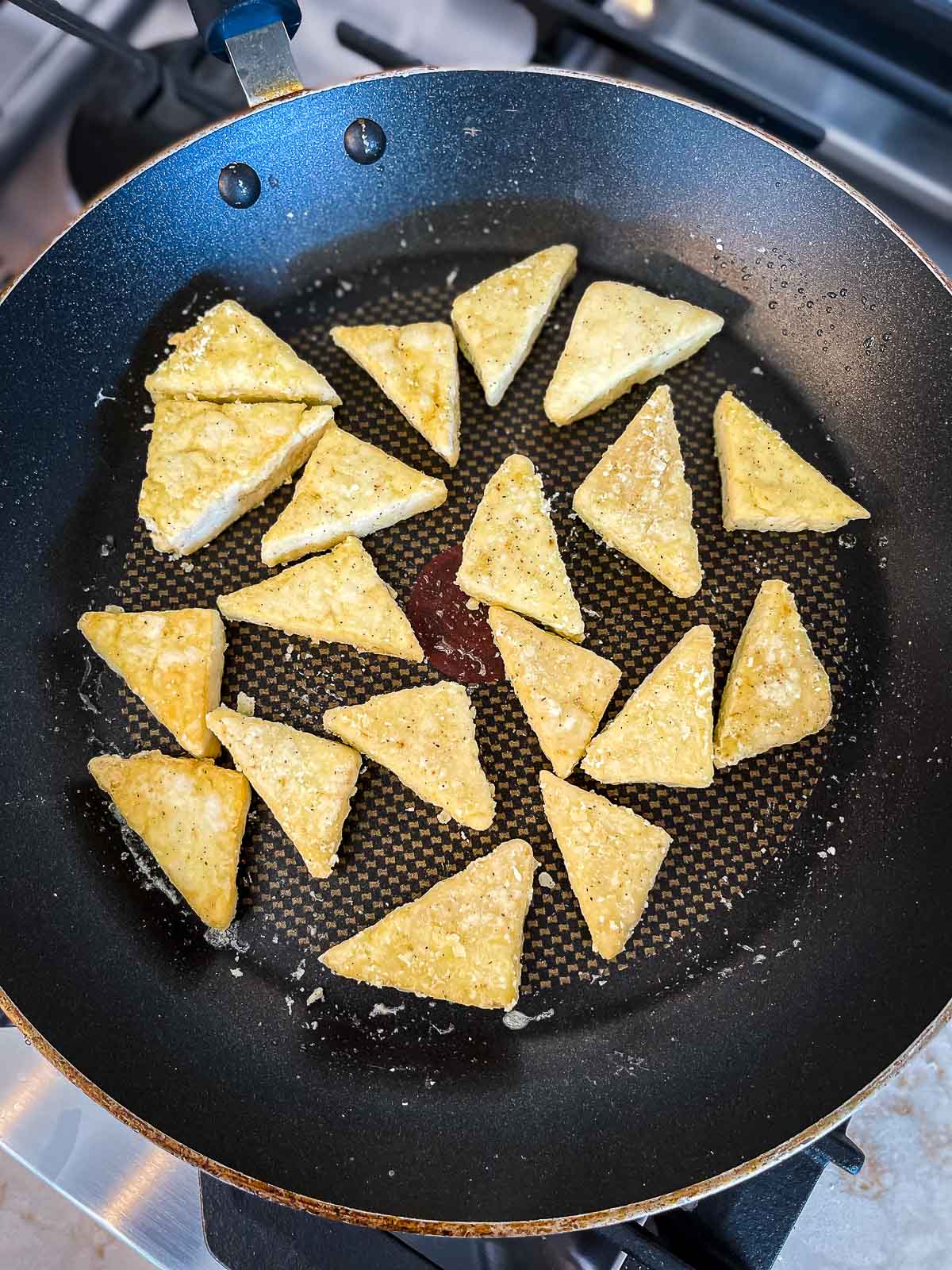 Tofu triangle steaks being fried and browned in a frying pan on top of a stove.
