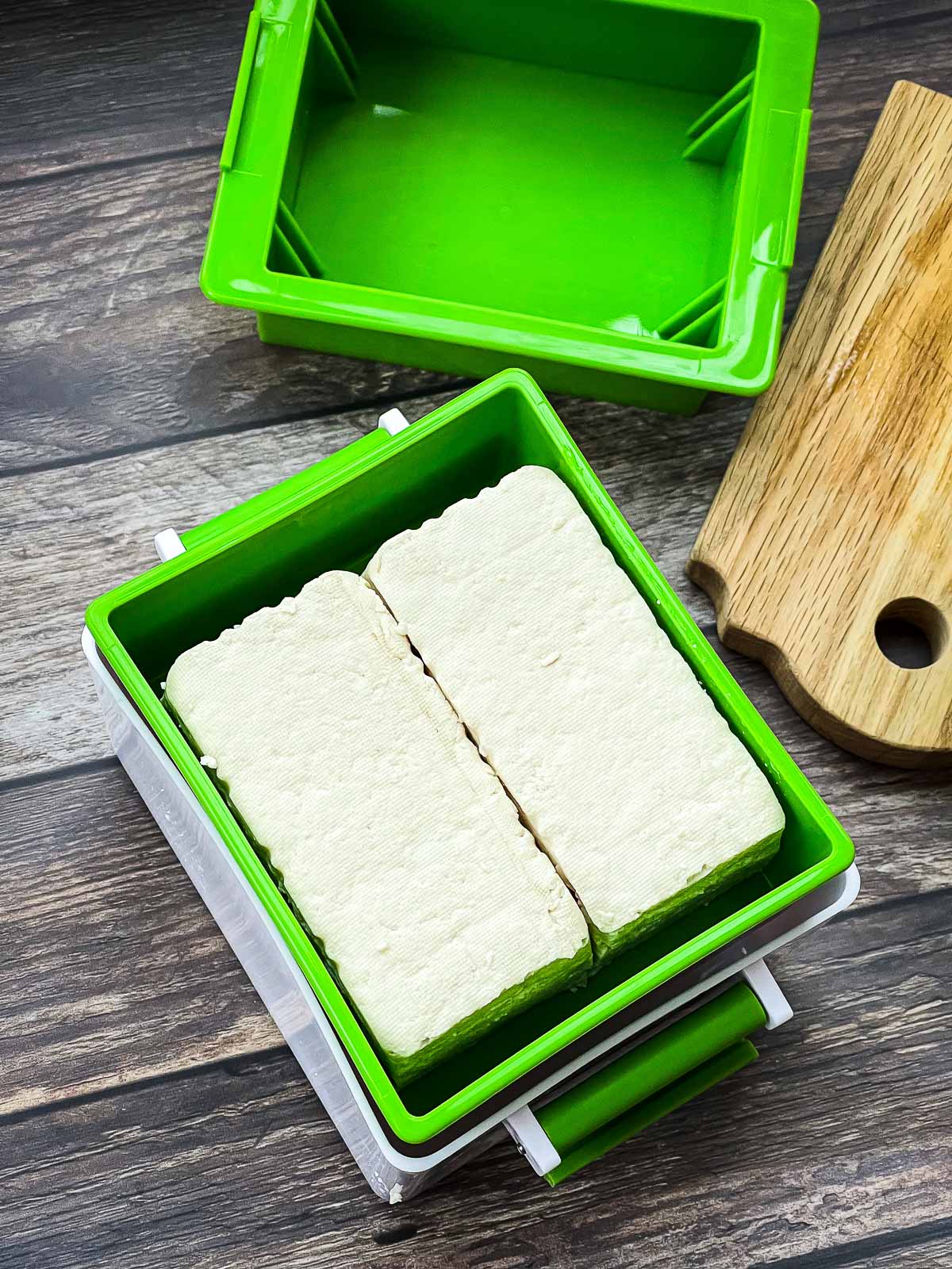 Tofu steaks inside of a green tofu press with a small wooden cutting board on the side.