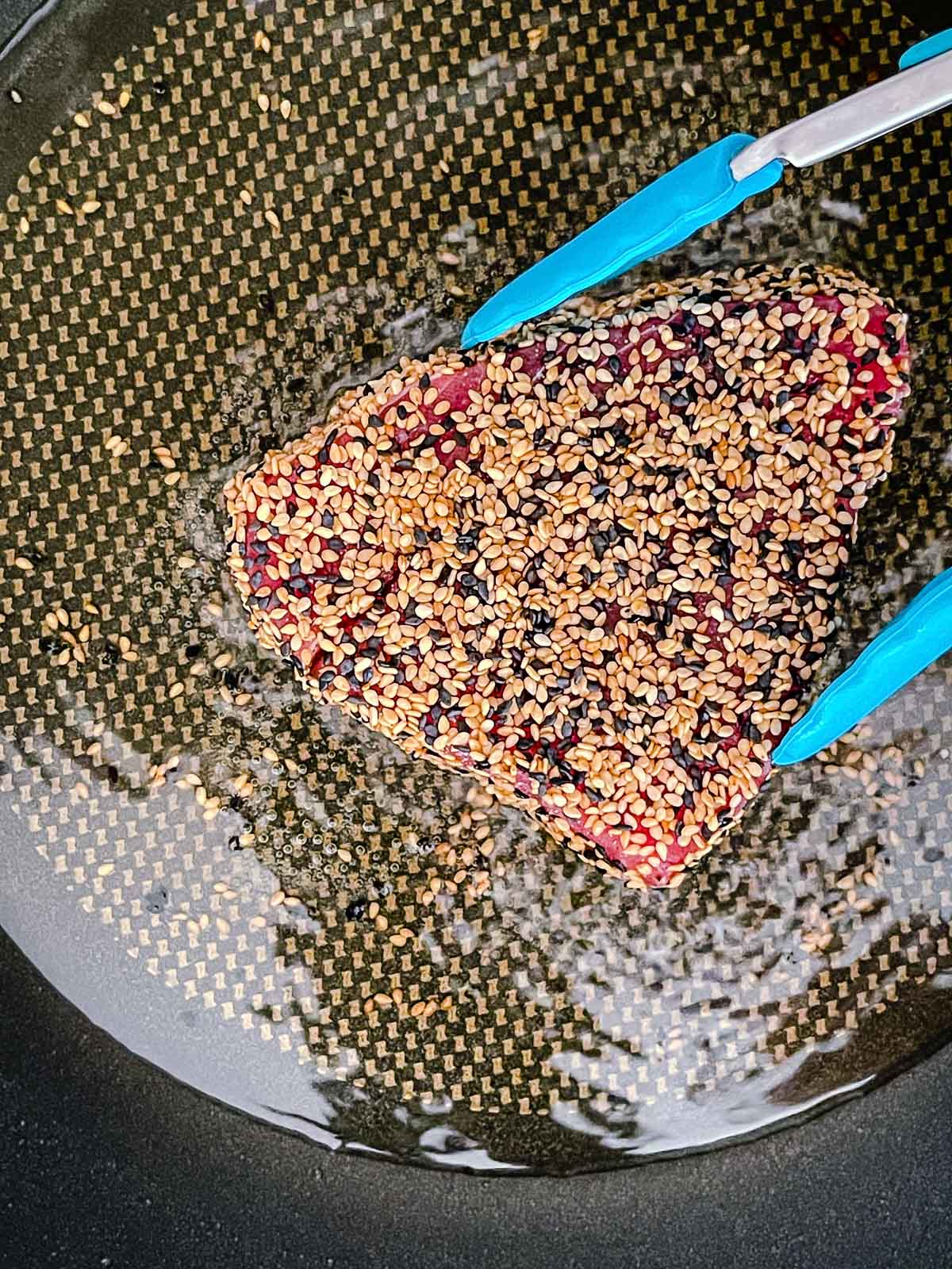 Blue tongs grabbing a tuna steak coated with sesame seeds that's been frying in a frying pan with oil.