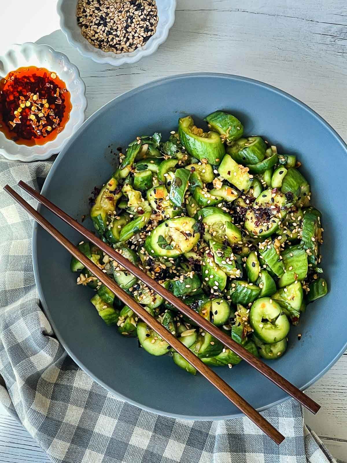Smashed cucumber salad in a gray bowl with wooden chopsticks on top and a small white bowl of chili oil, sesame seeds, and a checkered napkin on the side.