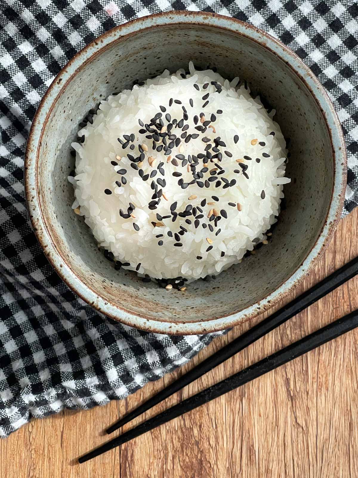 A gray bowl of steamed white rice sprinkled with black sesame seed on top of a checkered napkin with black chopsticks on the side.