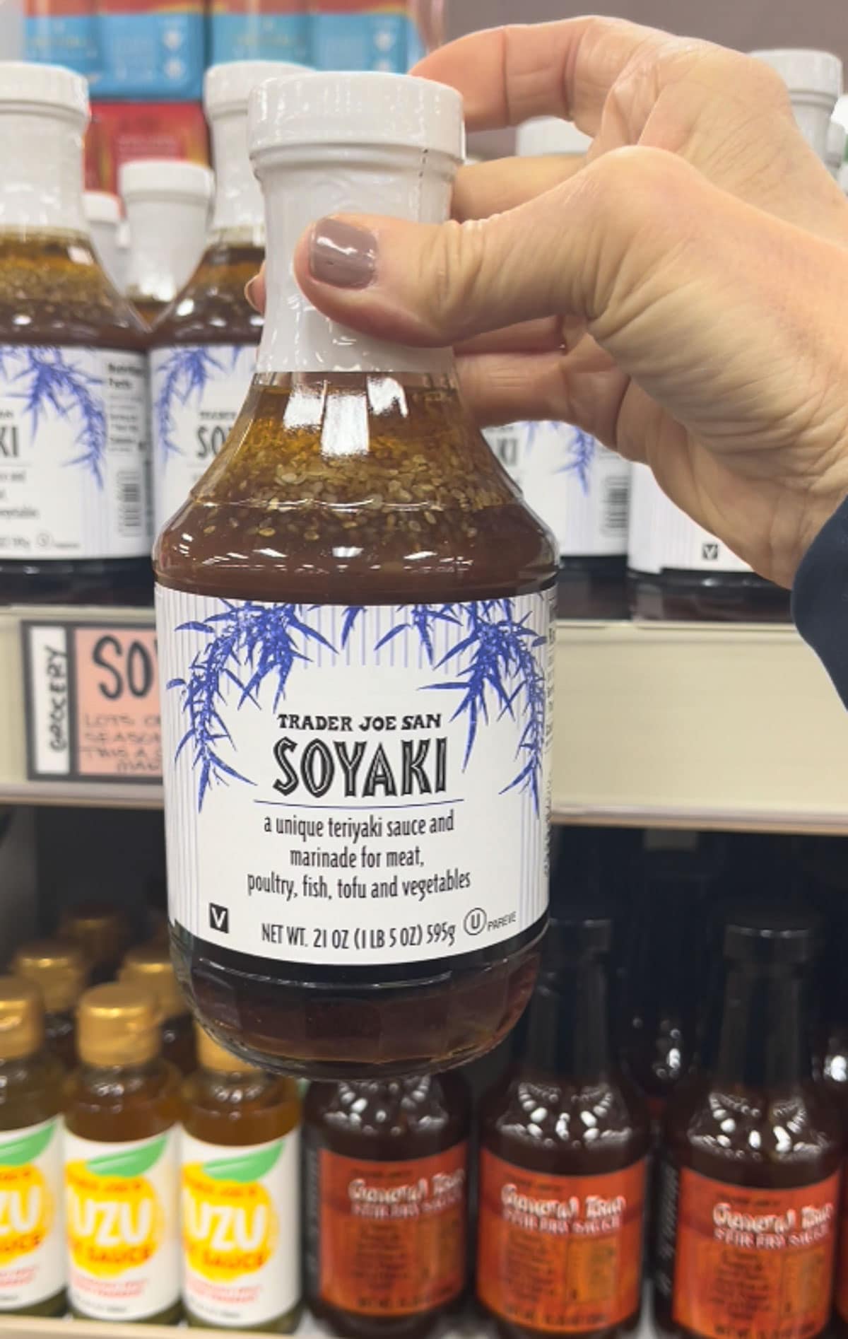 A hand holding up a bottle of Trader Joe's Soyaki teriyaki sauce with various bottles on shelves in the background.