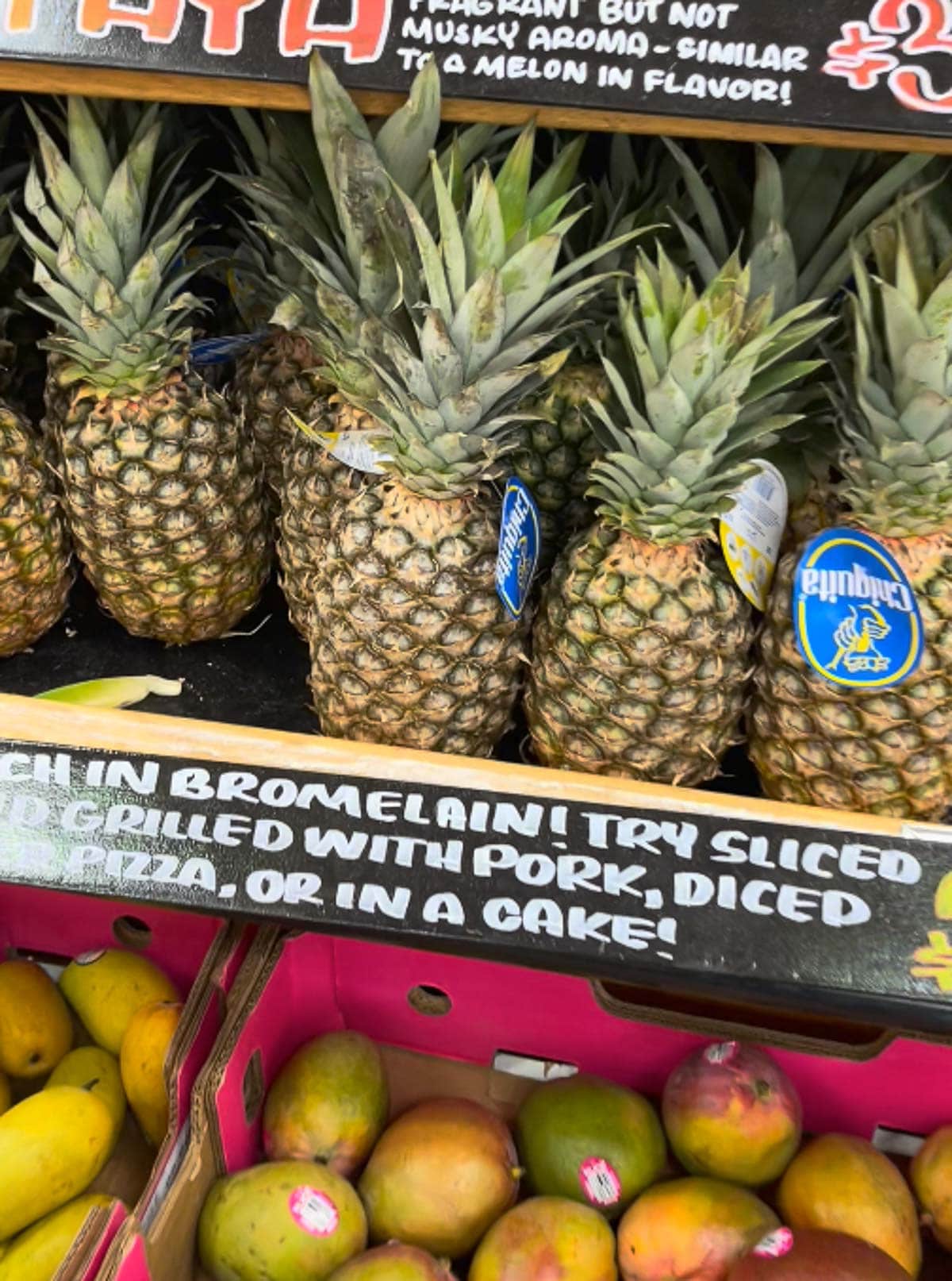 Fresh whole pineapples lined on a shelf at Trader Joe's with fresh mangos in boxes on a shelf below.