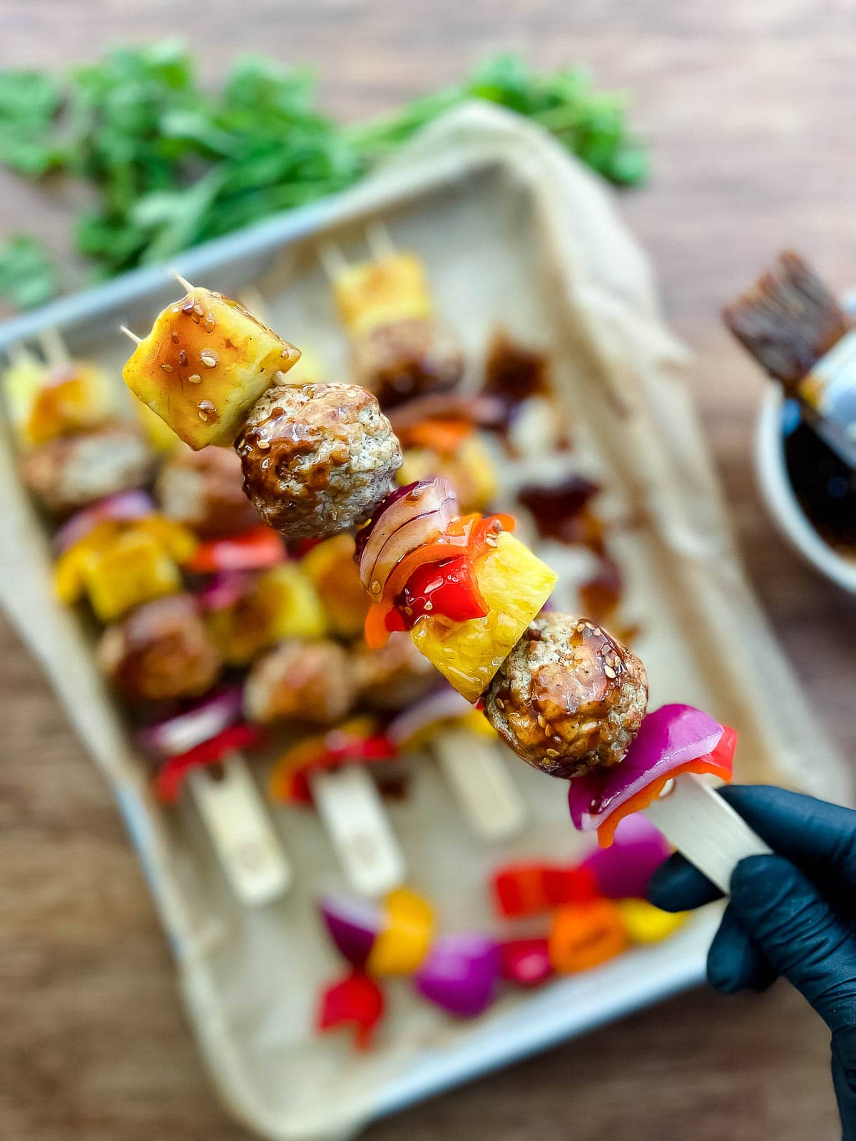 A close-up image of a gloved hand holding up a turkey meatball skewer ready for the grill, with other skewers beneath on top of a baking sheet.