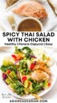 A white bowl filled with Spicy Thai Salad with Chicken with a fork on top placed on top of a marble surface, an a raw chicken breast and peanut dressing on parchment paper with a basting brush on the side..