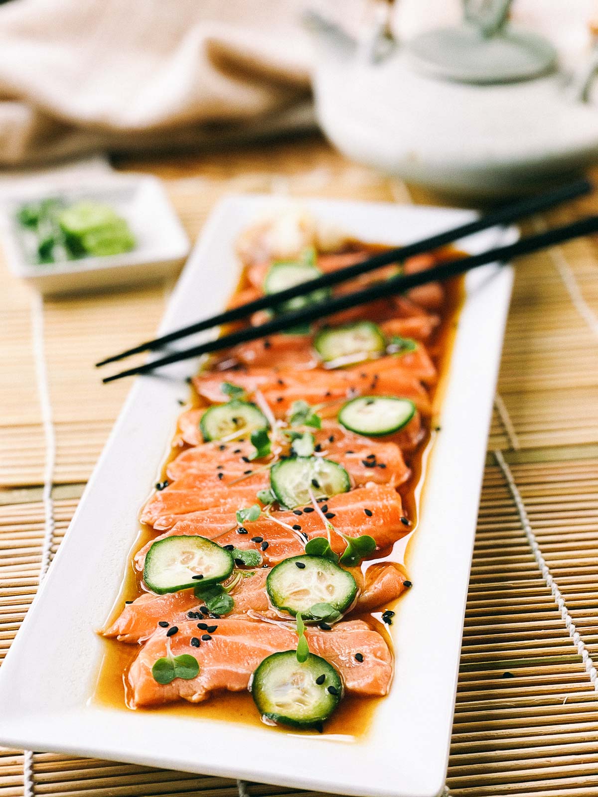 Slices of fresh sashimi salmon on a white platter with sliced cucumbers on top and sprinkles of black sesame seeds, with black chopsticks, a Japanese tea kettle, and a small white bowl of green wasabi on the side.