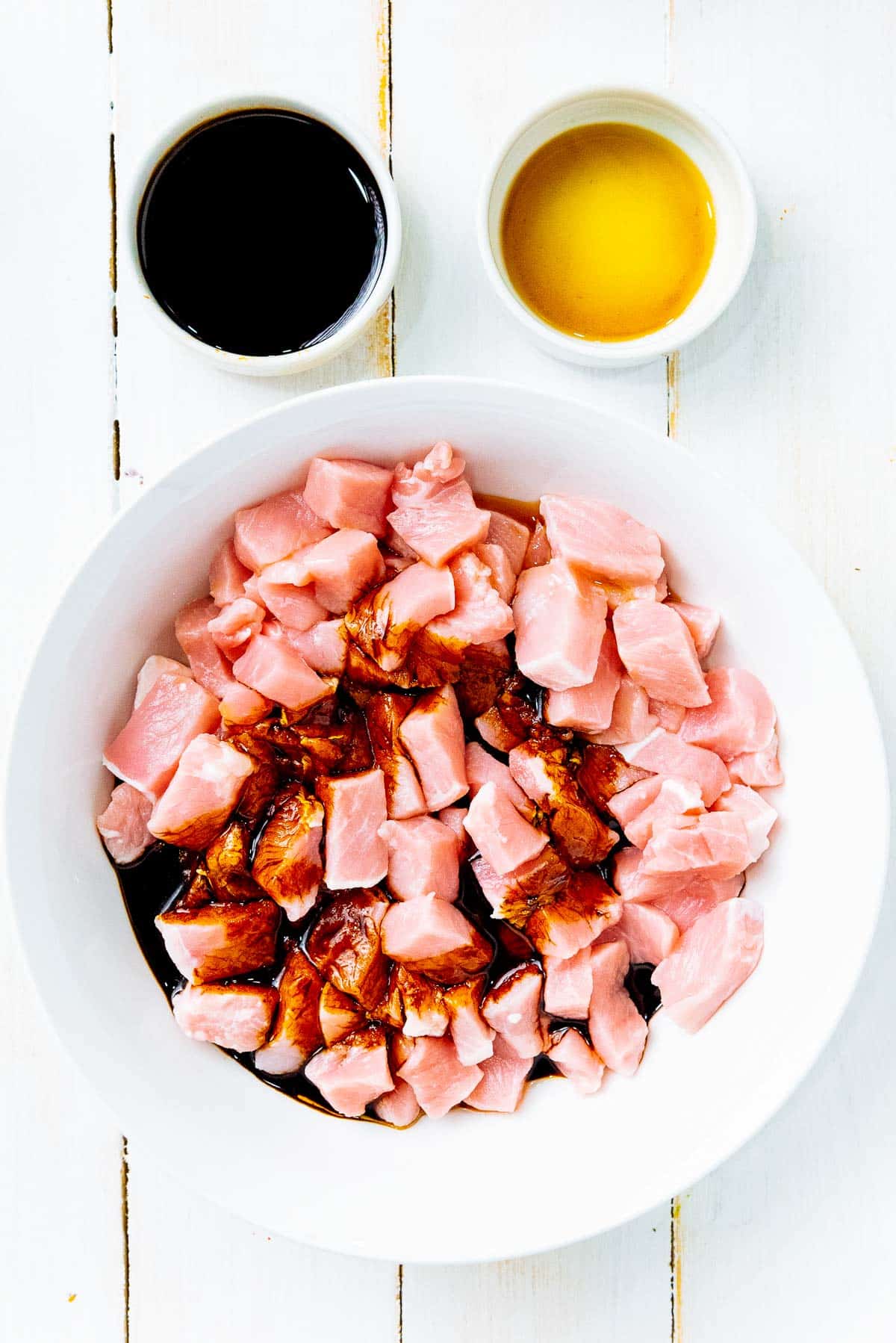 Pork chunks with marinade inside a white bowl with sauces in the background on top of a white board.
