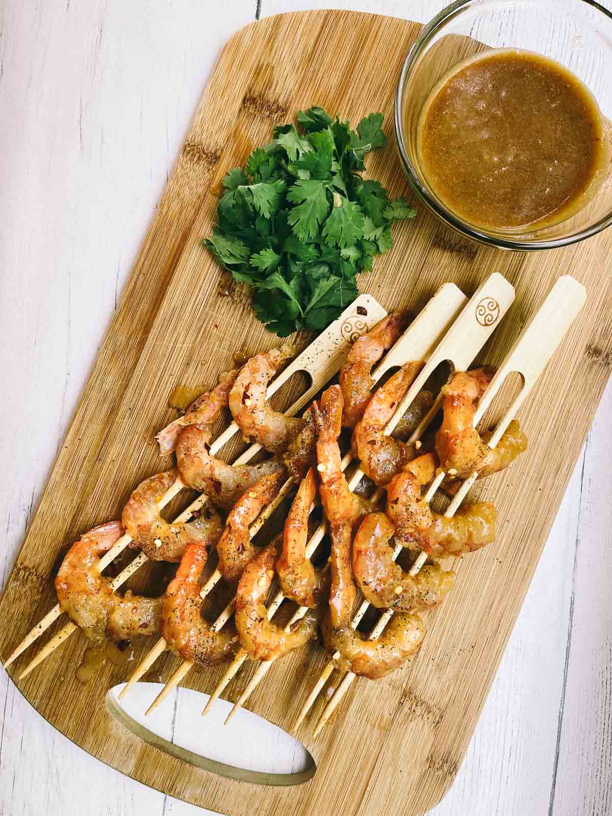 Shrimp on wooden skewers on top of a wooden cutting board ready to be grilled with a side of miso sauce and fresh cilantro.