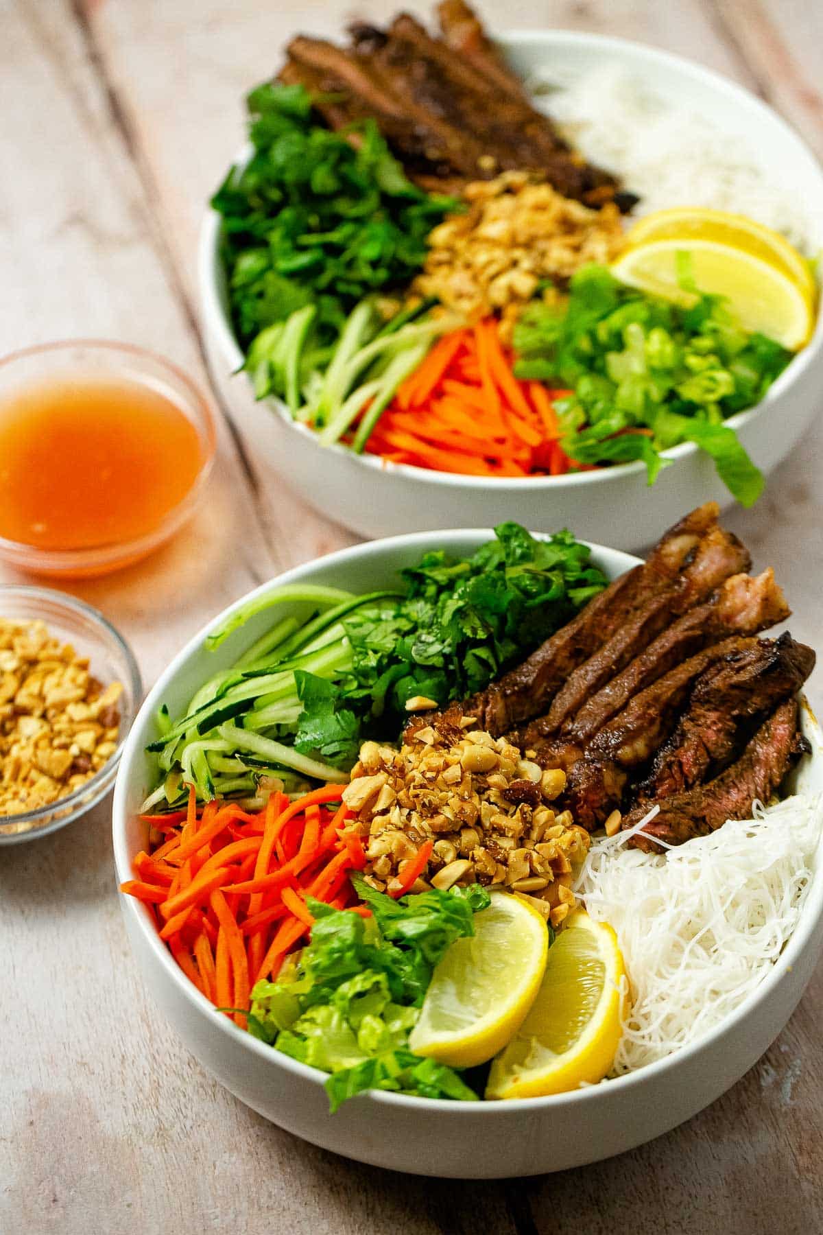 Asian Beef Noodle Bowls in two large white bowls with chopped peanuts and a sauce on the side on top of a wooden board.