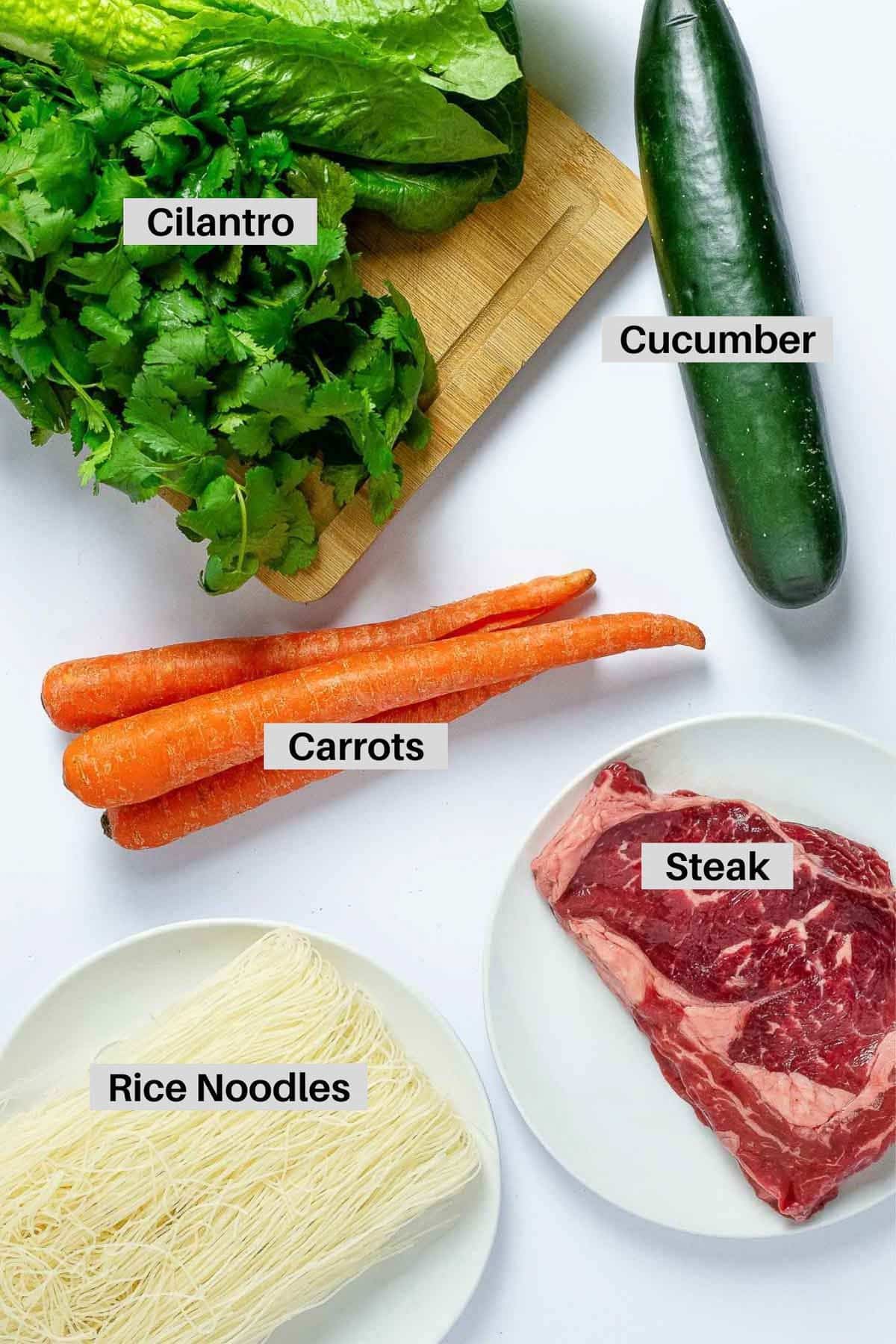 Labeled ingredients in bowls and on a board for making Asian beef noodle bowls on top of a white surface.