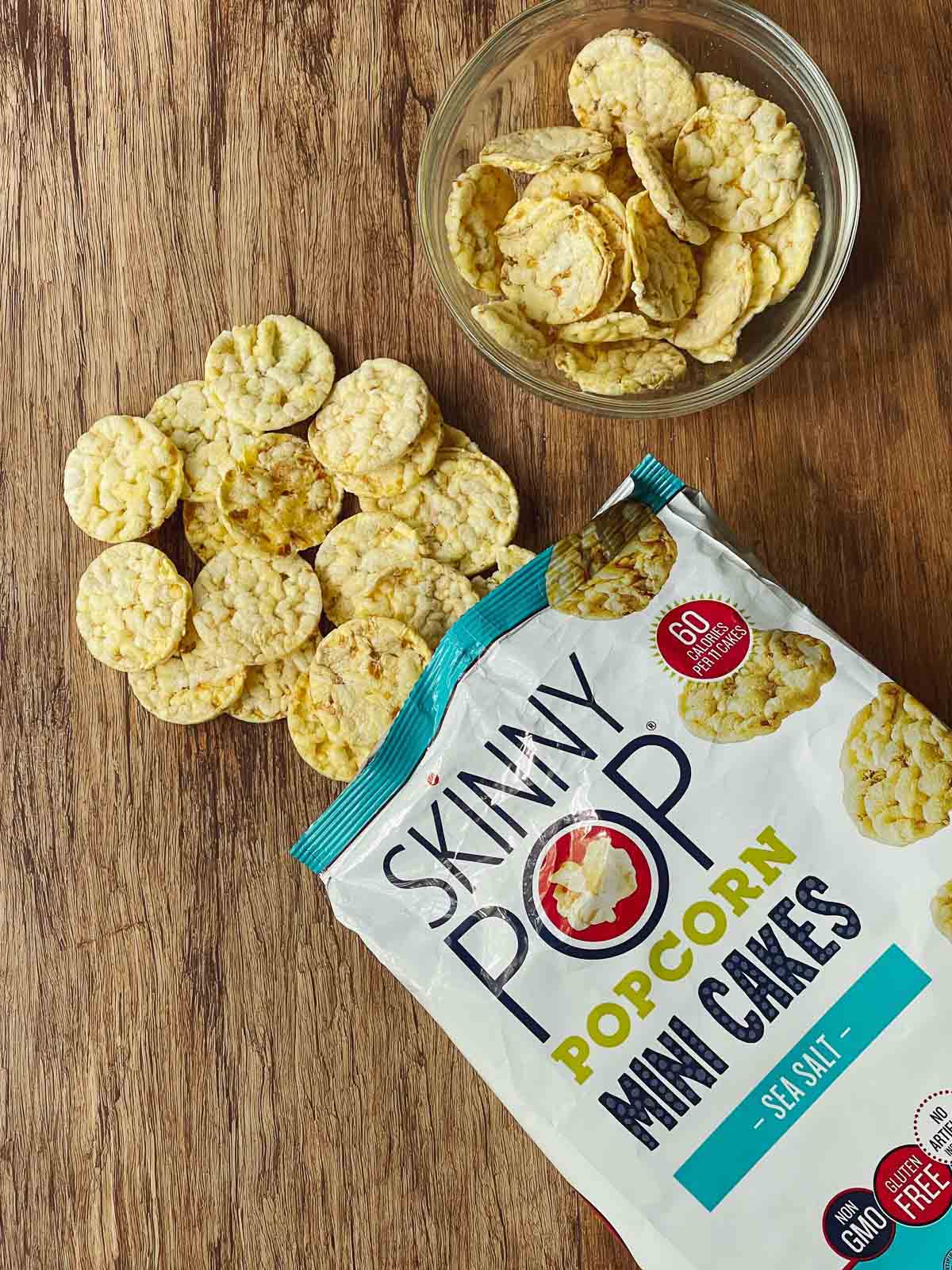 A bag of Skinny Pop mini popcorn cakes on top of a wooden board with mini cakes coming out of the bag and a side of mini cakes in a glass bowl.