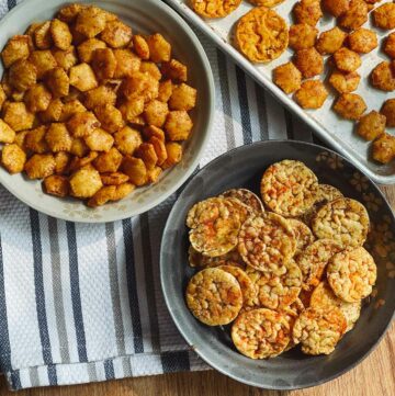 Sriracha popcorn cakes in a gray bowl and Sriracha oyster crackers in a light gray bowl on top of a striped kitchen towel and a baking tray with more crackers on the side.