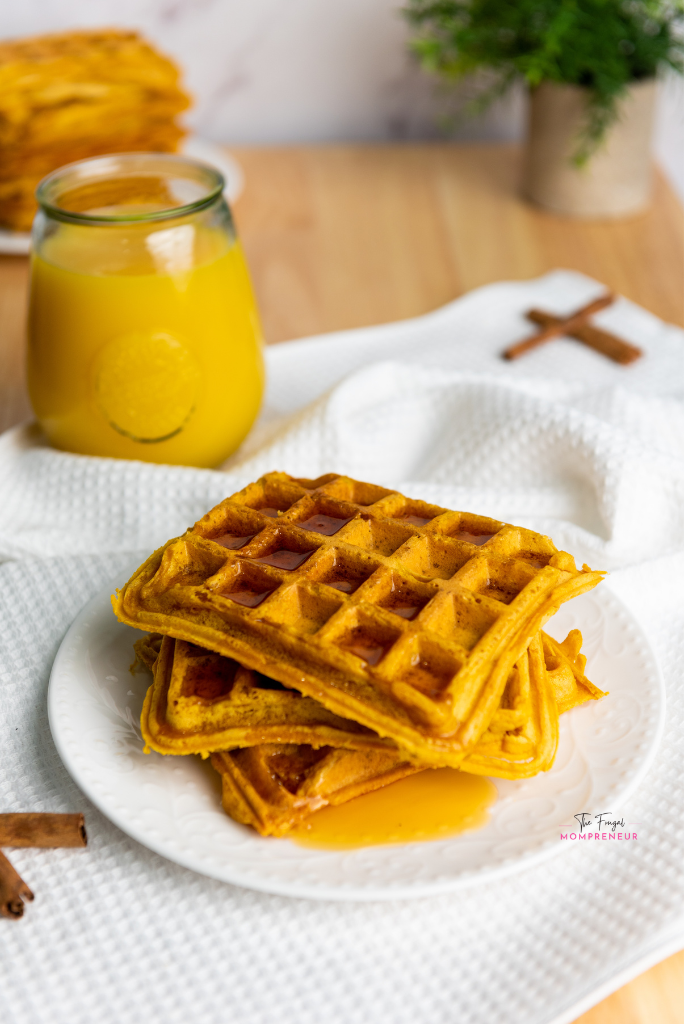 A stack of vegan pumpkin waffles on a white plate with a glass of orange juice in the background.