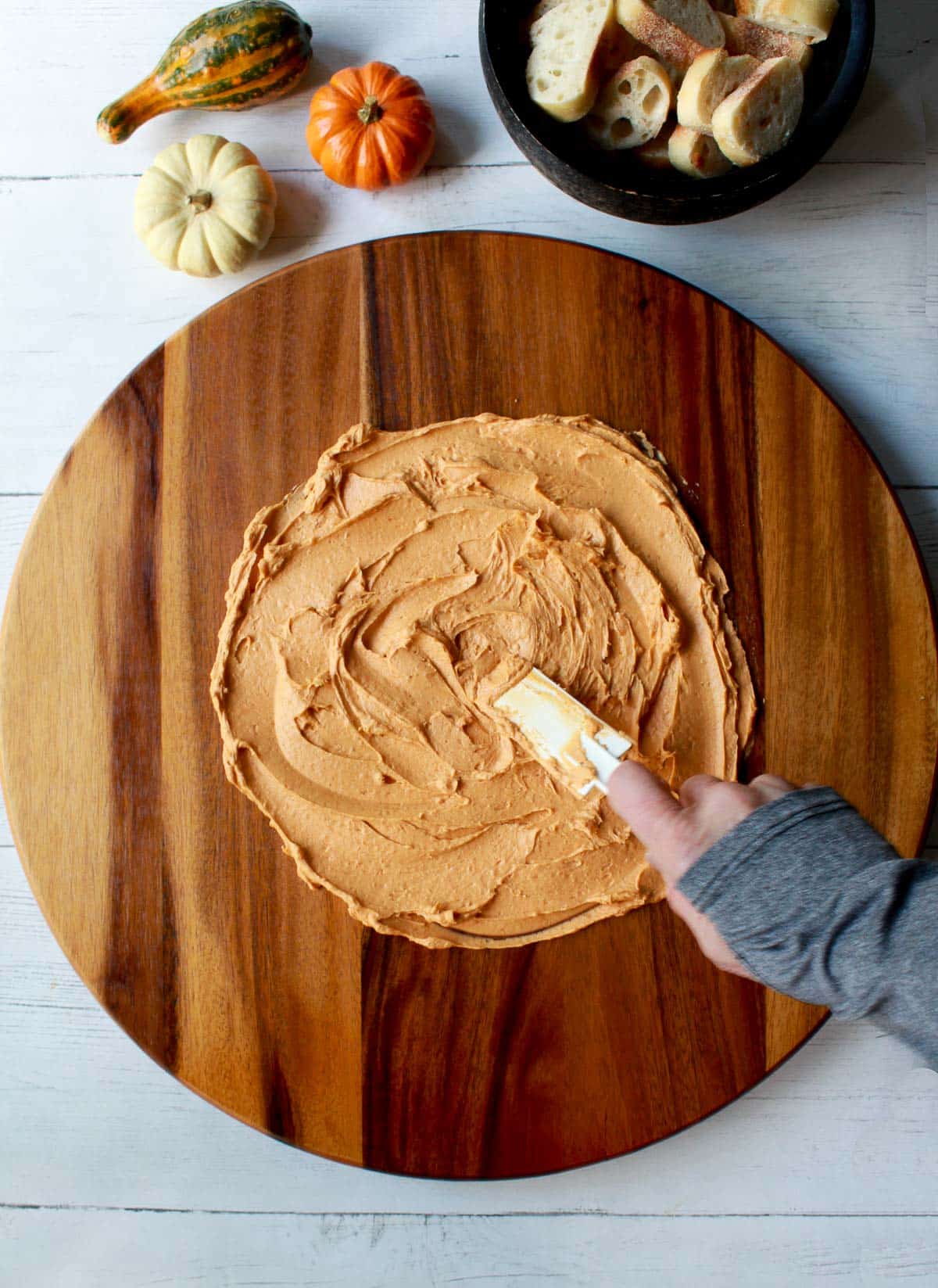 a hand spreading miso butter onto a round wooden board with baby pumpkins and a bowl of bread slices in the background