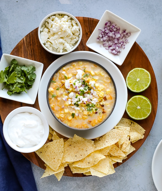 A bowl of creamy street corn soup on a round wooden board surrounded by toppings in white bowls and tortilla chips and limes on the side.