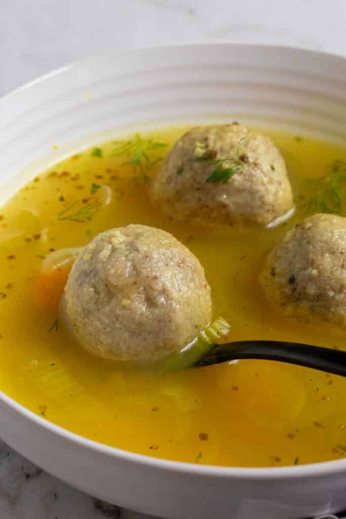 A white bowl of vegan matzo ball soup with a spoon lifting one of the matzo balls.