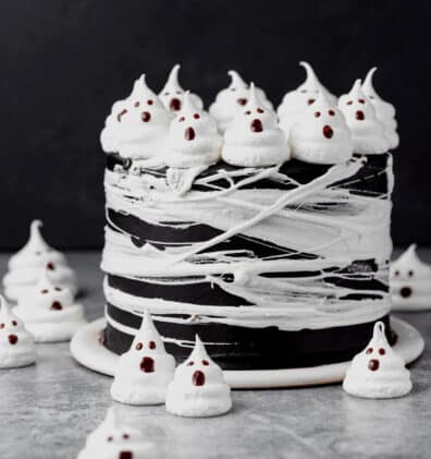 A black, layered Halloween cocoa ghost cake topped with meringue ghosts and wrapped with marshmallow spider webs on top of a gray surface.