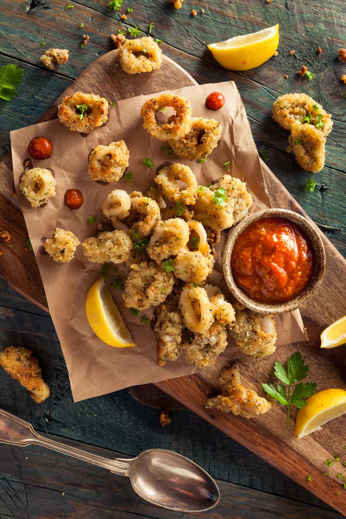 Crispy, fried calamari on parchment paper on top of a wooden board with a side of dipping sauce and lemon wedges.