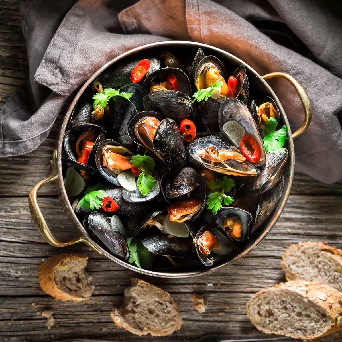 An overhead shot of a copper pot filled with steamed mussels garnished with red chili peppers and cilantro on a wooden board with sliced bread and a gray napkin on the side.