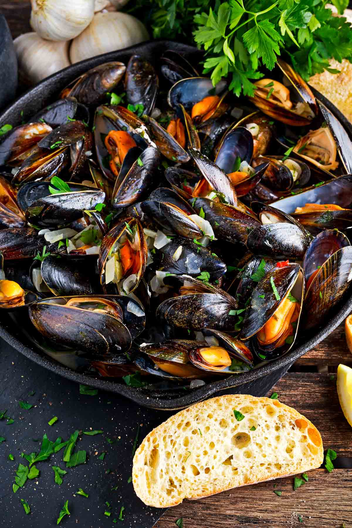 A pot filled with Thai curry steamed mussels garnished with red chili peppers and cilantro on a wooden board with sliced bread, garlic, and green herbs on the side.