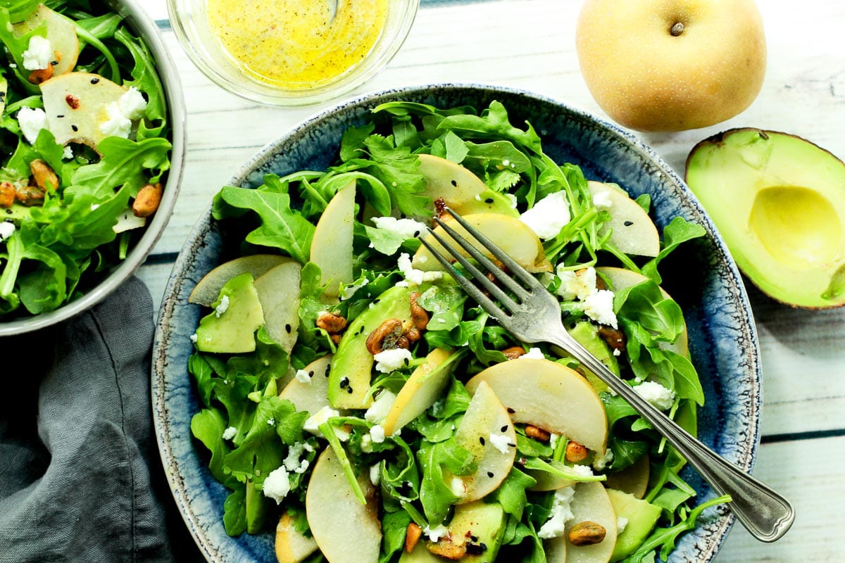 An arugula salad with sliced Asian pears and avocado in a blue bowl with a fork on top and half an avocado, whole pear, and lemon vinaigrette dressing on the side.