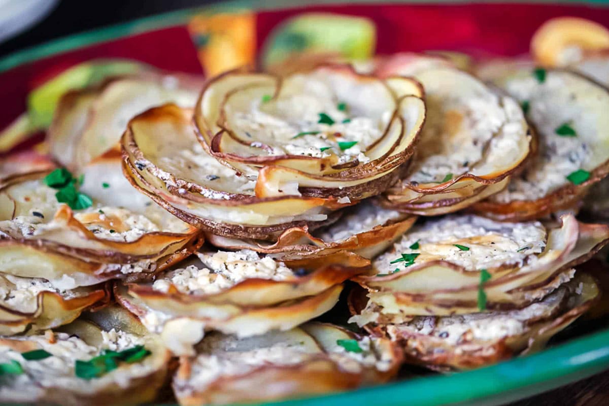 Thin slices of potatoes stacks holiday side dish stacked high on a green platter ad topped with Boursin cheese and fresh herbs.