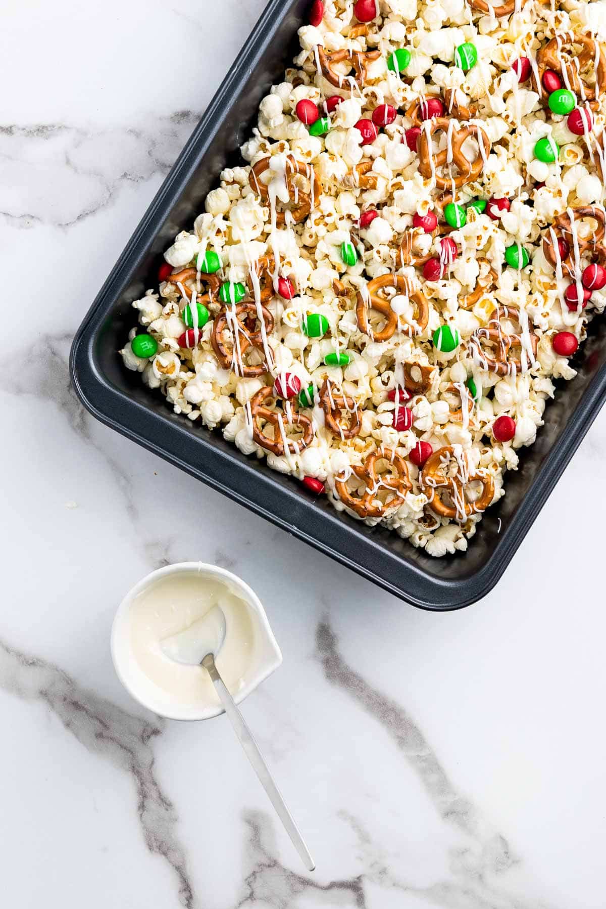 Christmas popcorn Santa munch drizzled with white chocolate in a black baking tray with white chocolate on the side on top of a marble surface.