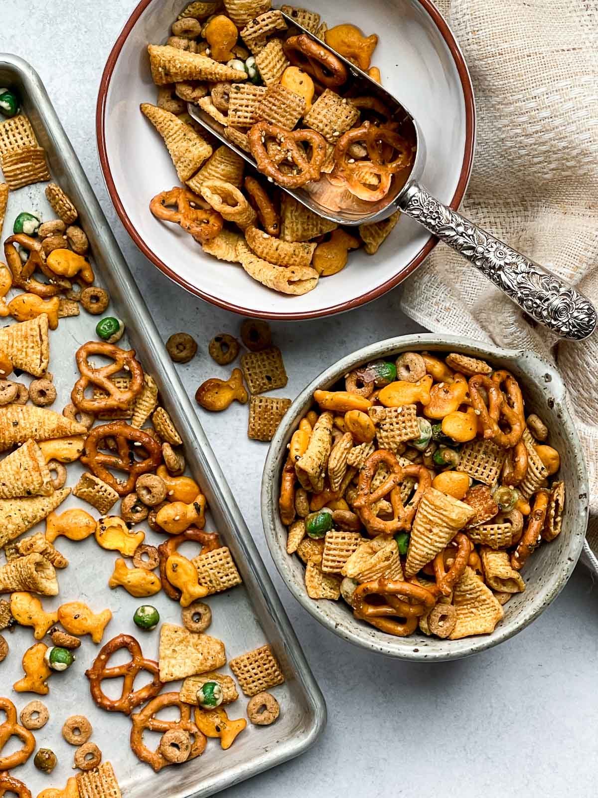 Two bowls of Furikake Chex Mix, one with a silver scoop, on a gray surface with a linen napkin and baking tray of Chex Mix on the side.
