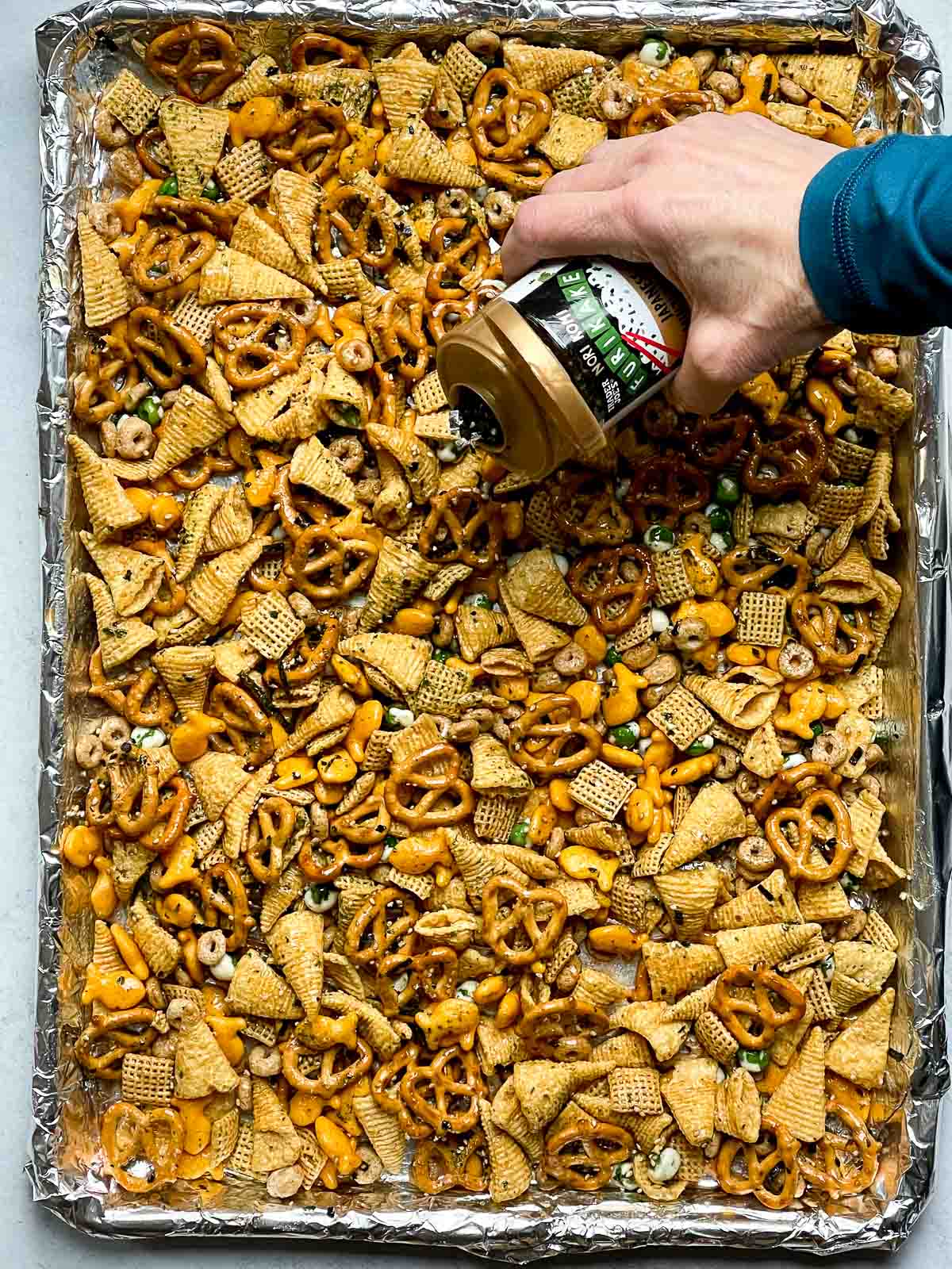 A baking tray lined with Furikake Chex Mix with a hand adding the Furikake seasoning.