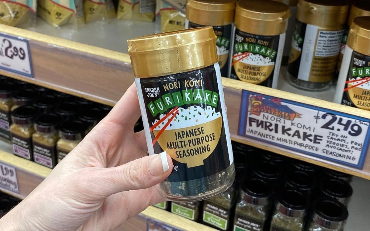 A bottle of Trader Joe's Nori Komi Furikake seasoning being held up by a woman's hand in front of a spice shelf.
