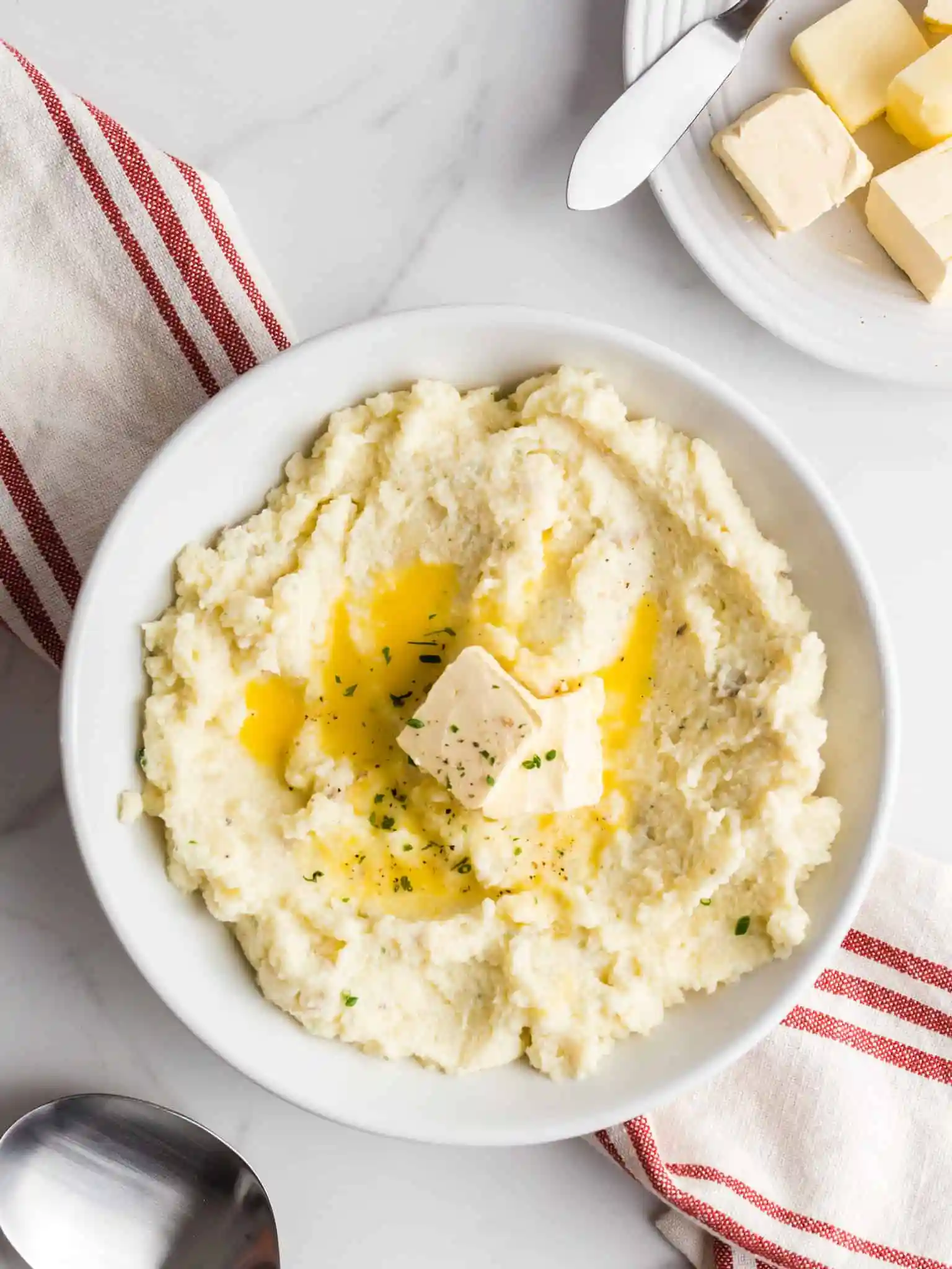 A round white bowl with garlic mashed potatoes topped with fresh herbs and melted butter, with a small plate of butter slices on the side on top of a red and white napkin.