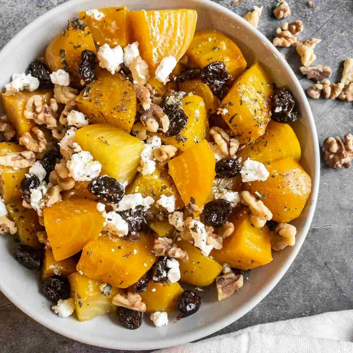 Chunks of roasted golden beets in a round white bowl tossed with roasted cherries, walnuts, and pieces of goat cheese.