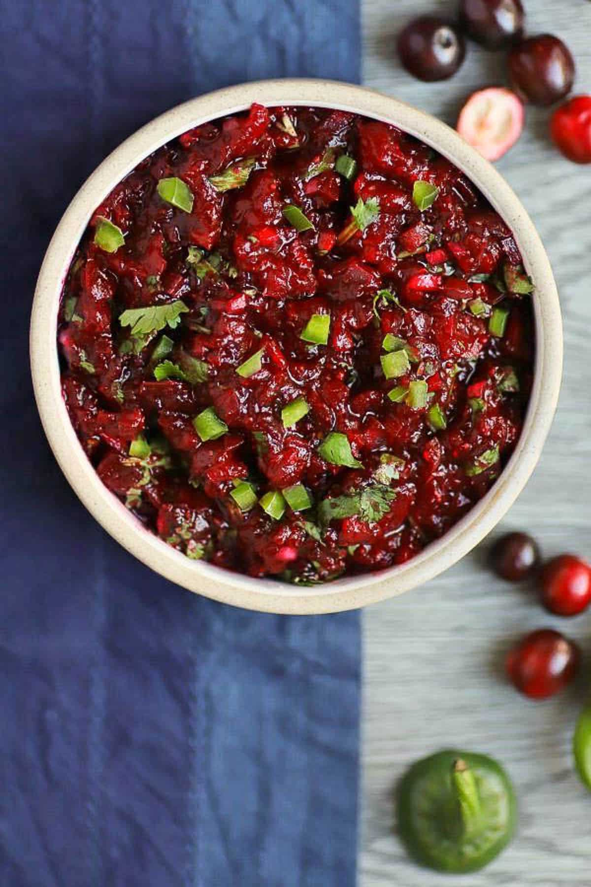 A large round white bowl filled with jalapeño cranberry salsa holiday side dish topped with herbs on top of a blue napkin with fresh cranberries on the side.