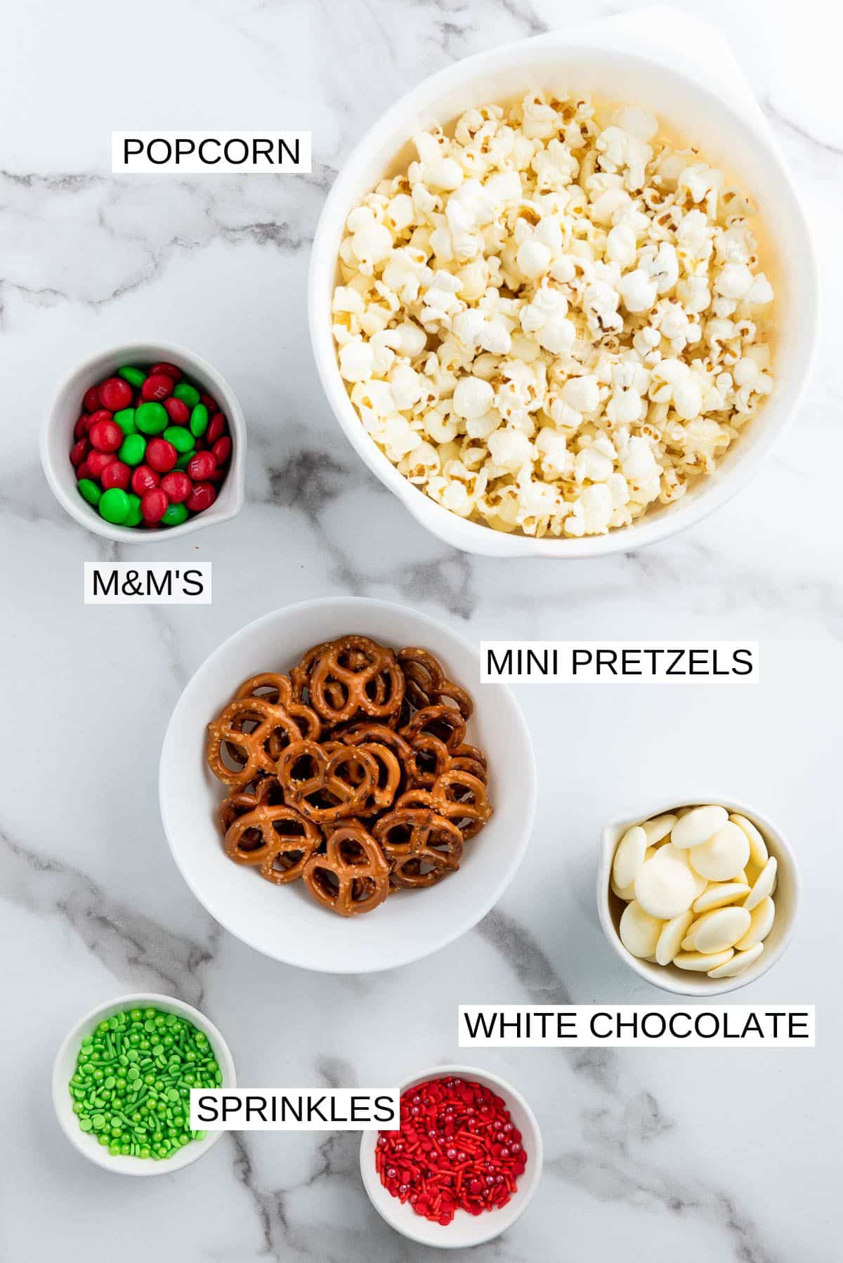 Labeled ingredients in white bowls for making Christmas Popcorn Santa Munch on a marble surface.