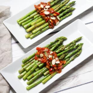 Roasted asparagus spears on a long white plates topped with a sweet and spicy pepper sauce on top of a white wooden board.