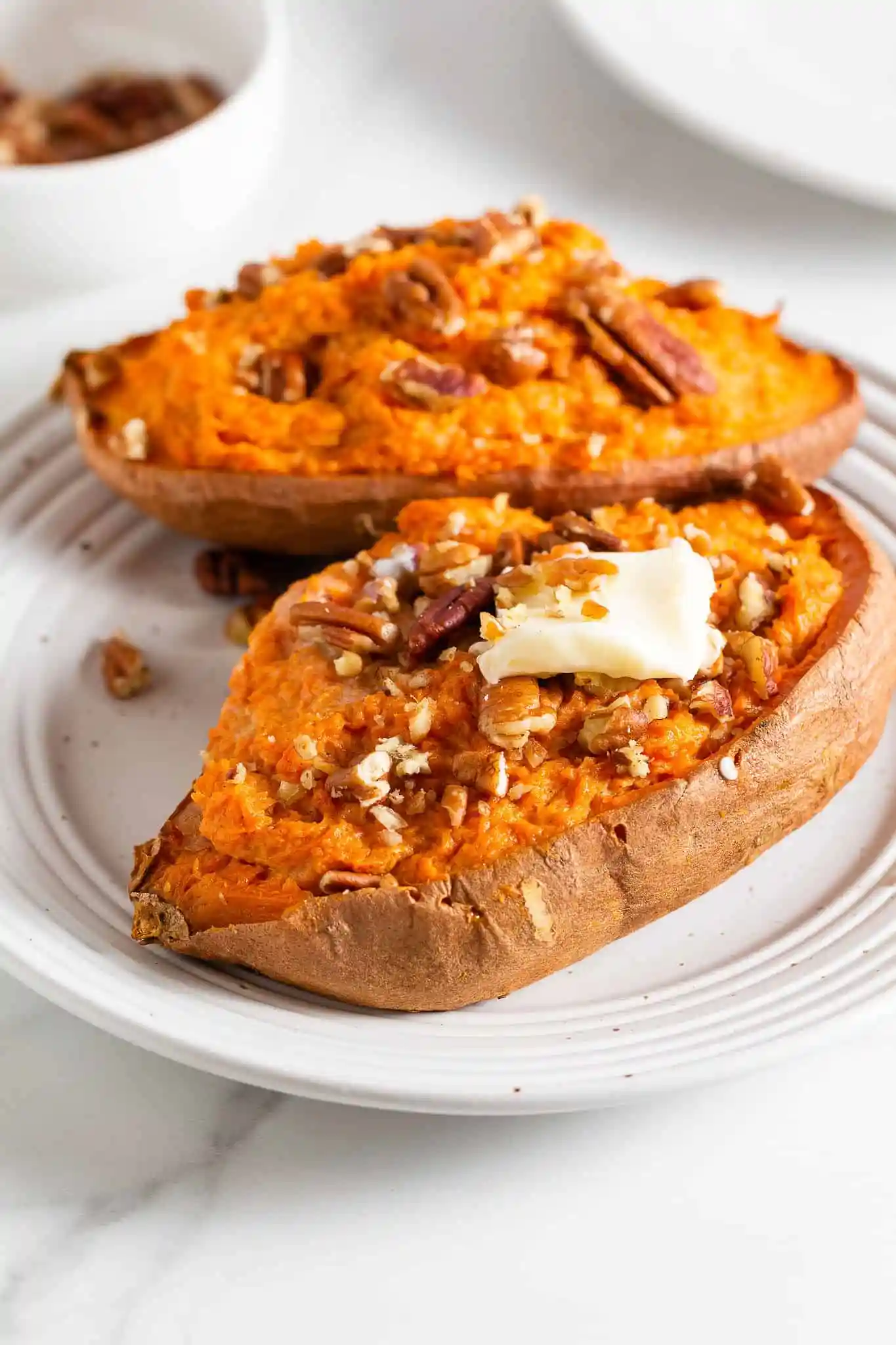 Two roasted sweet potato halves holiday side dish topped with pecans and a slice of melted butter on top of a round white plate.