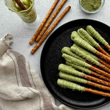 Matcha chocolate dipped pretzel rods on top of a round black plate with plain pretzel rods on the side, and a small bowl of matcha powder and a glass with a dipped pretzel rod, and a linen napkin on the side.