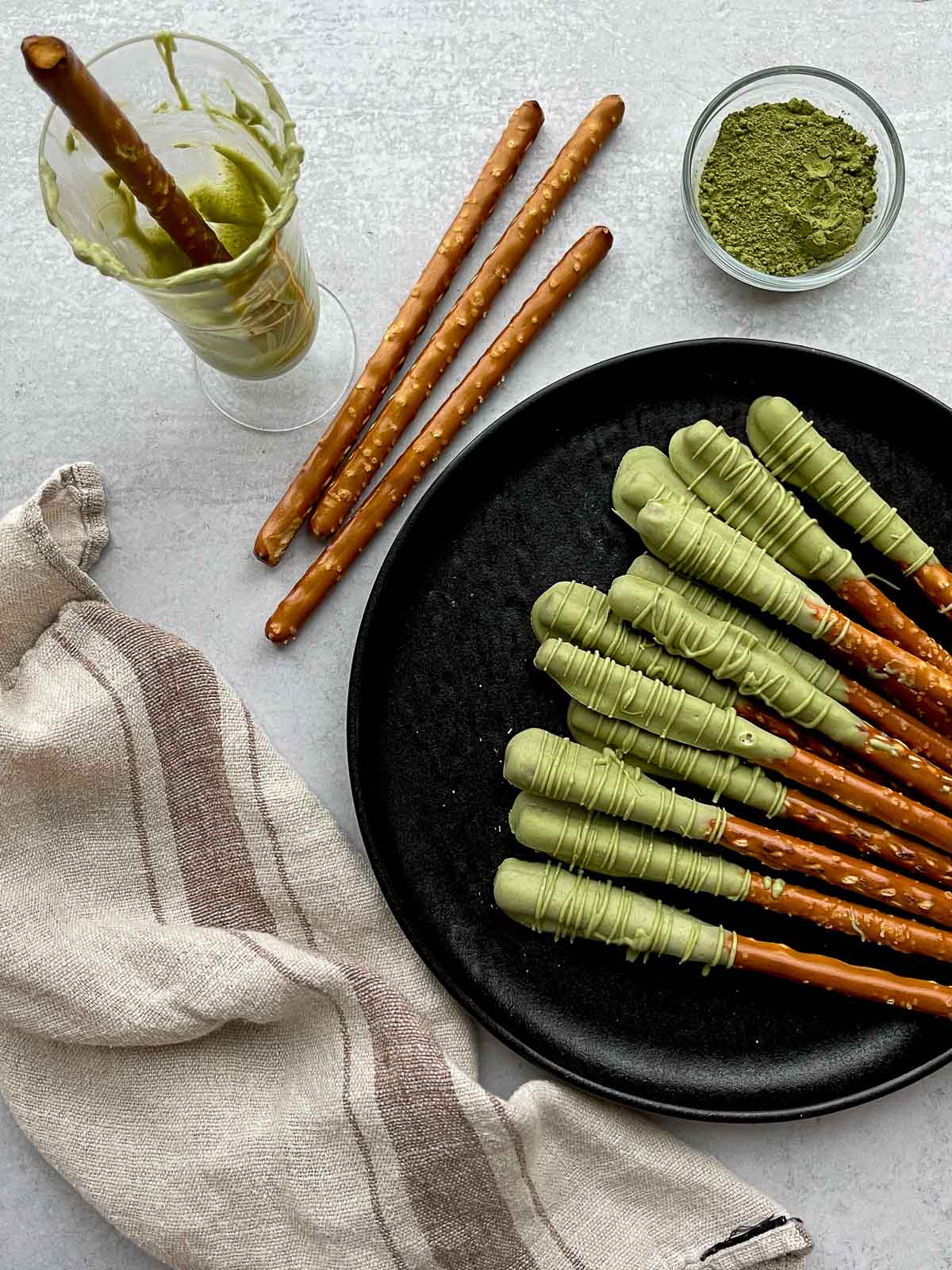 Matcha chocolate dipped pretzel rods on top of a round black plate with plain pretzel rods on the side, and a small bowl of matcha powder and a glass with a dipped pretzel rod, and a linen napkin on the side.