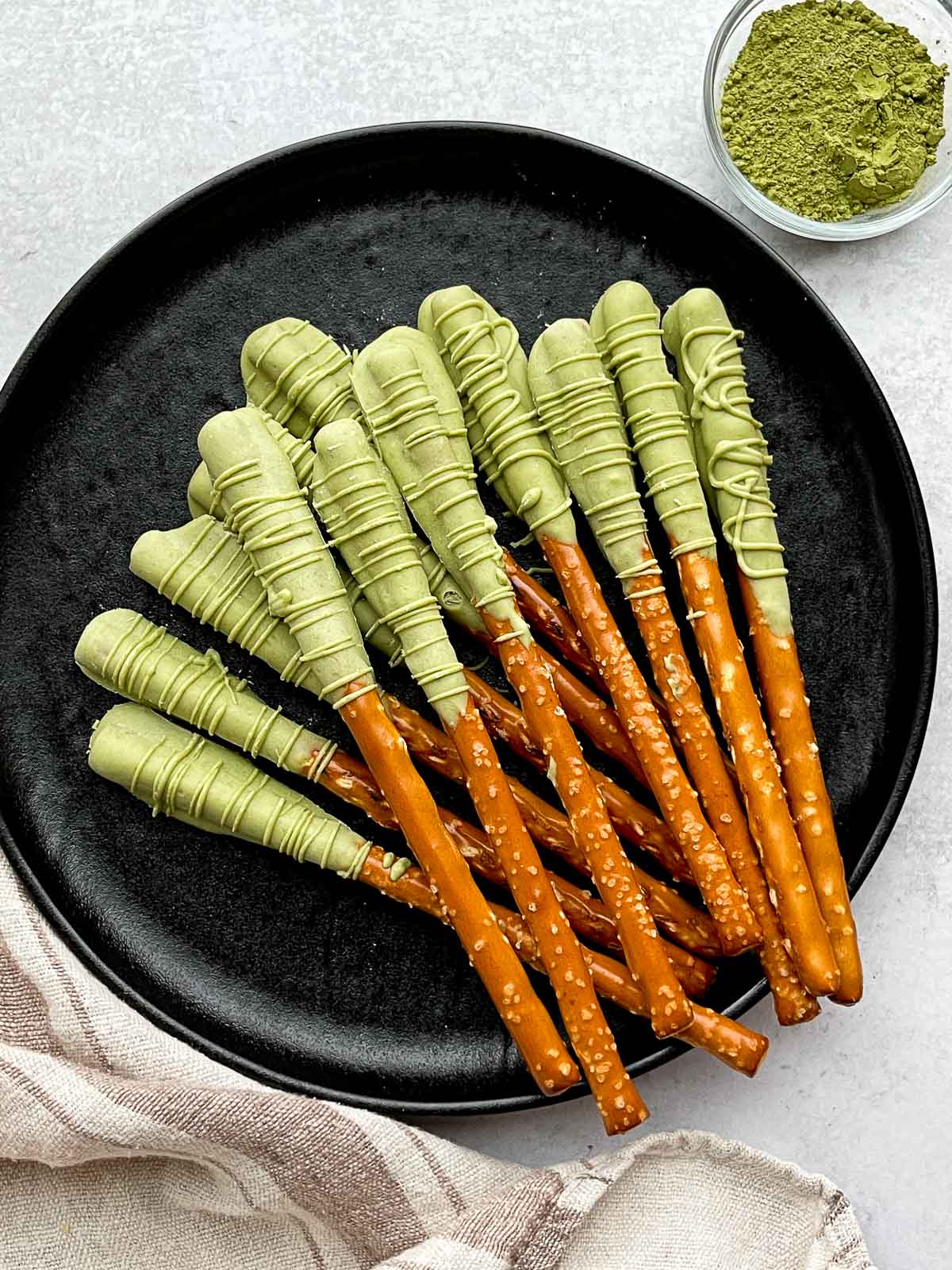 Matcha chocolate dipped pretzel rods on top of a round black plate with plain pretzel rods on the side, and a small bowl of matcha powder and a linen napkin on the side.