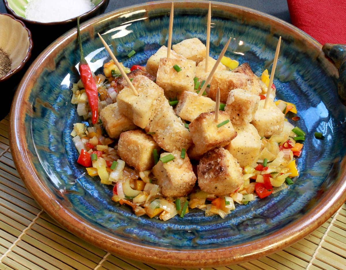 Crispy salt and pepper tofu cubes piled on top of sauteed vegetables on a round blue plate with a small bowl of salt on the side.