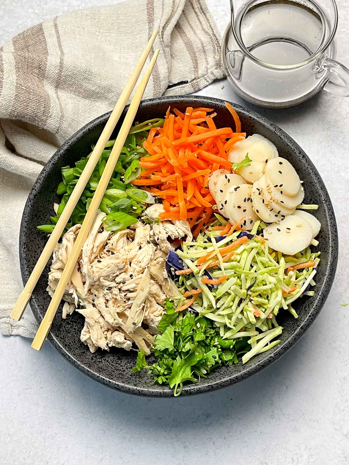 A large black bowl filled with Chinese chicken salad featuring shredded chicken, shredded carrots, broccoli slaw, scallions, water chestnuts and chow mein noodles, with a pair of chopsticks on top and dressing in a glass pourer, and a linen napkin on the side.