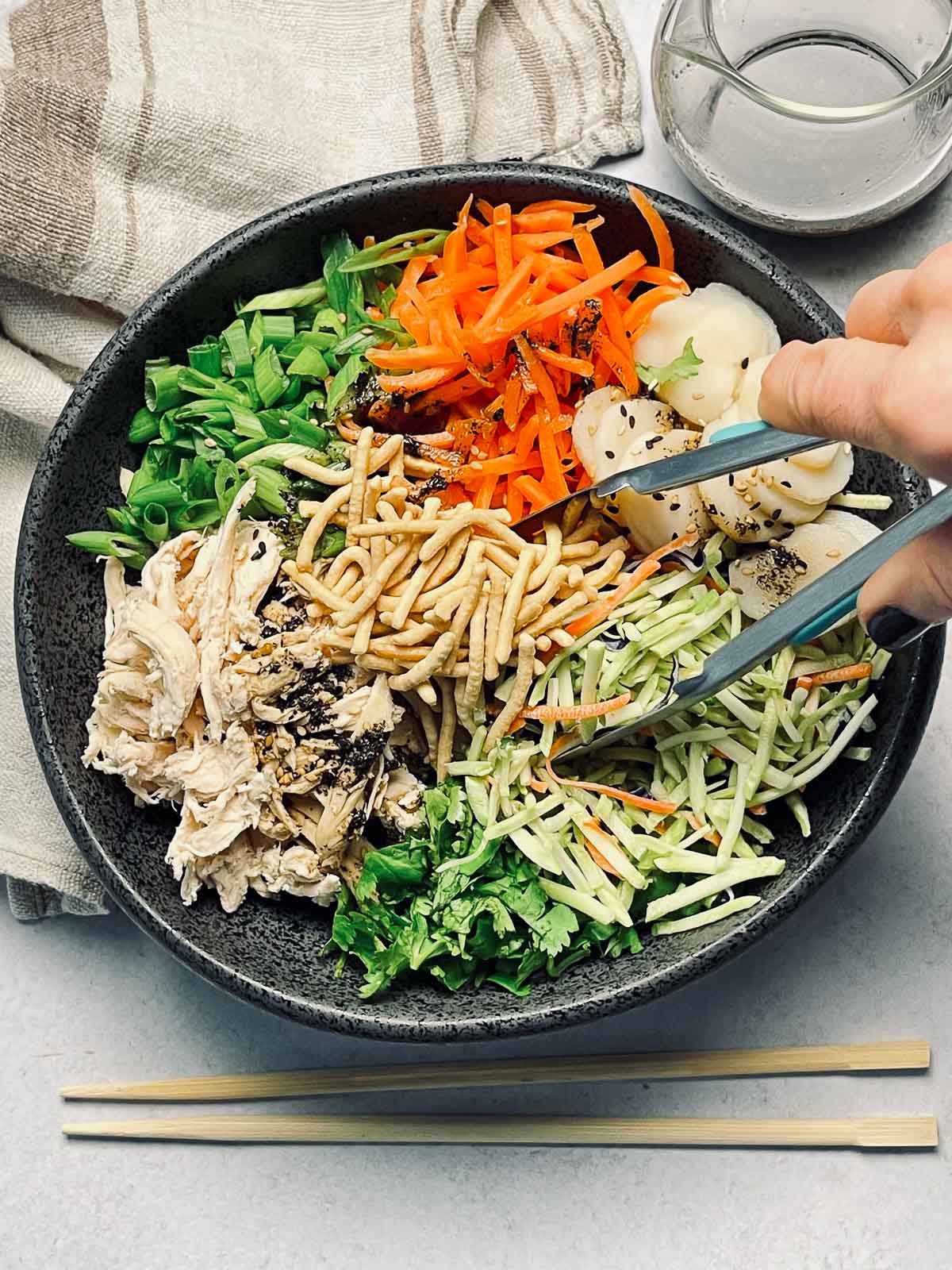 A hand with tongs with large black bowl filled with Chinese chicken salad featuring shredded chicken, shredded carrots, broccoli slaw, scallions, water chestnuts and chow mein noodles, with a pair of chopsticks, a linen napkin, and dressing in a glass pourer on the side.