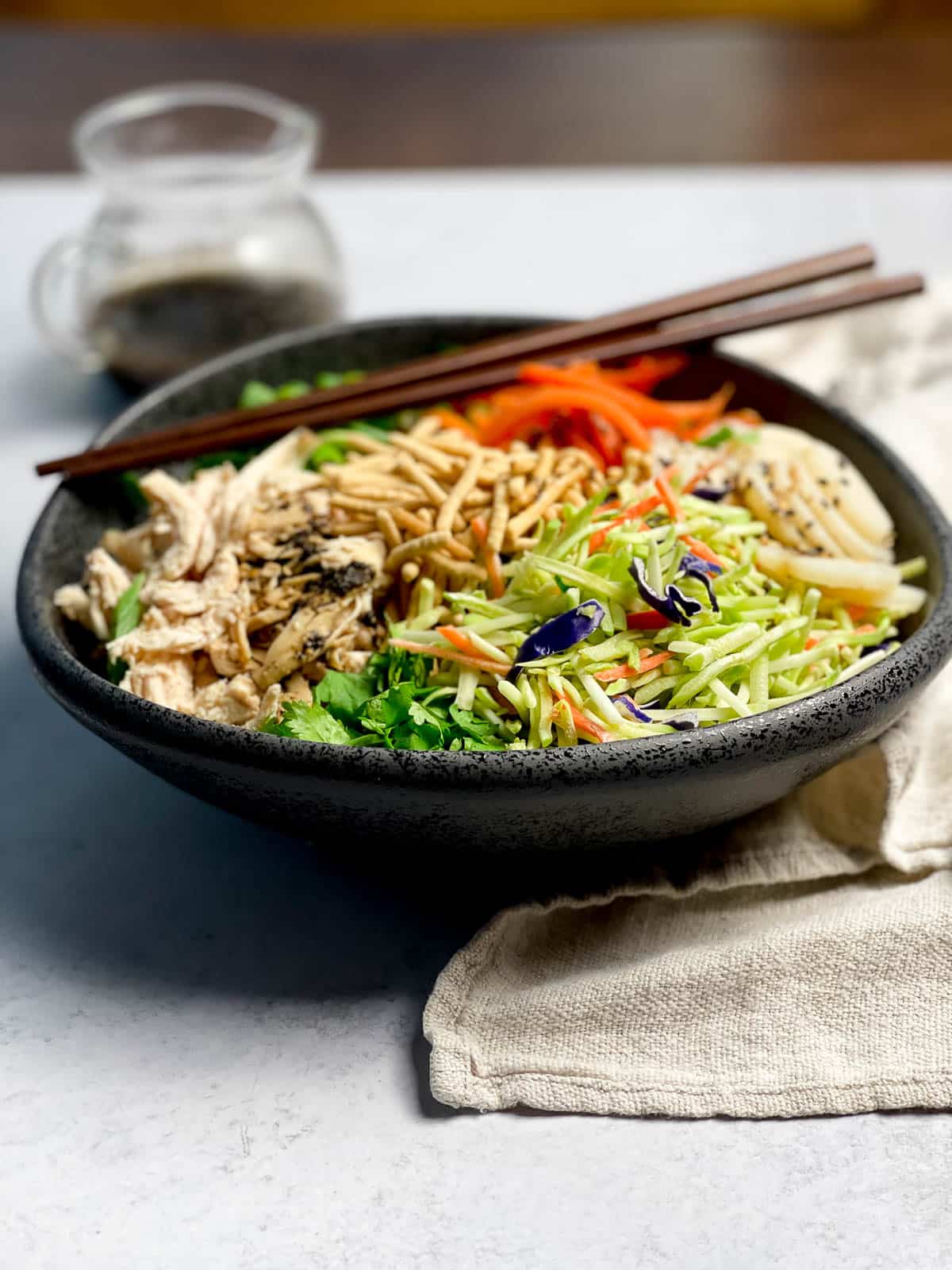 A large black bowl filled with Chinese chicken salad featuring shredded chicken, shredded carrots, broccoli slaw, scallions, water chestnuts and chow mein noodles, with a pair of brown chopsticks on top and a linen napkin on the side.