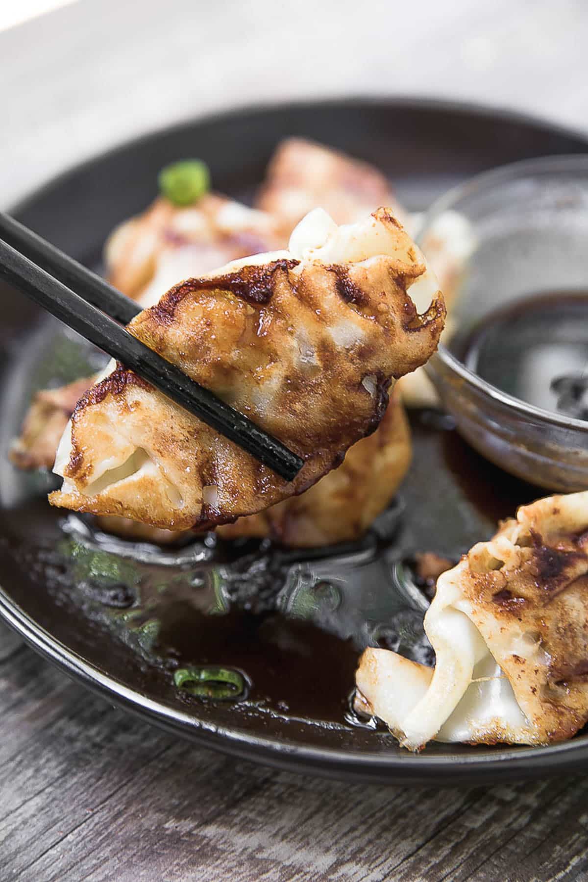Pork Potstickers held by chopsticks on a black plate with a clear glass bowl of dipping sauce on a dark wooden board