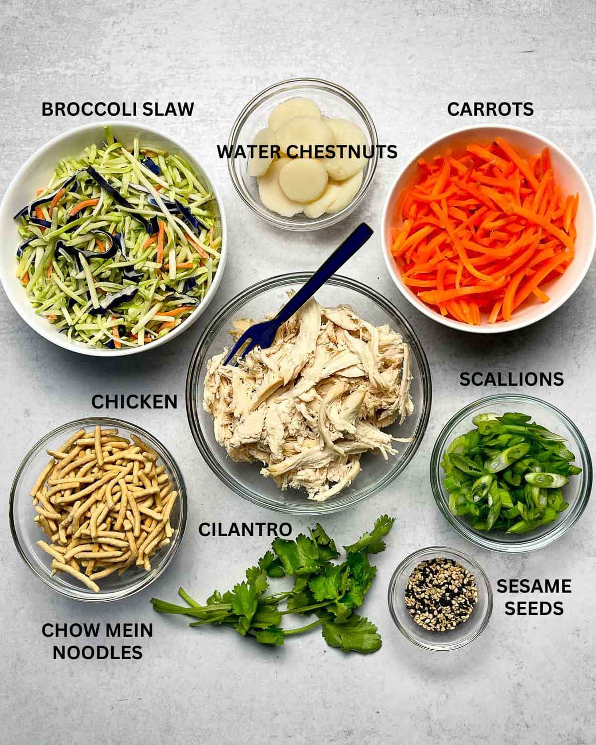 Labeled ingredients in small bowls for making Chinese chicken salad on top of a gray surface.