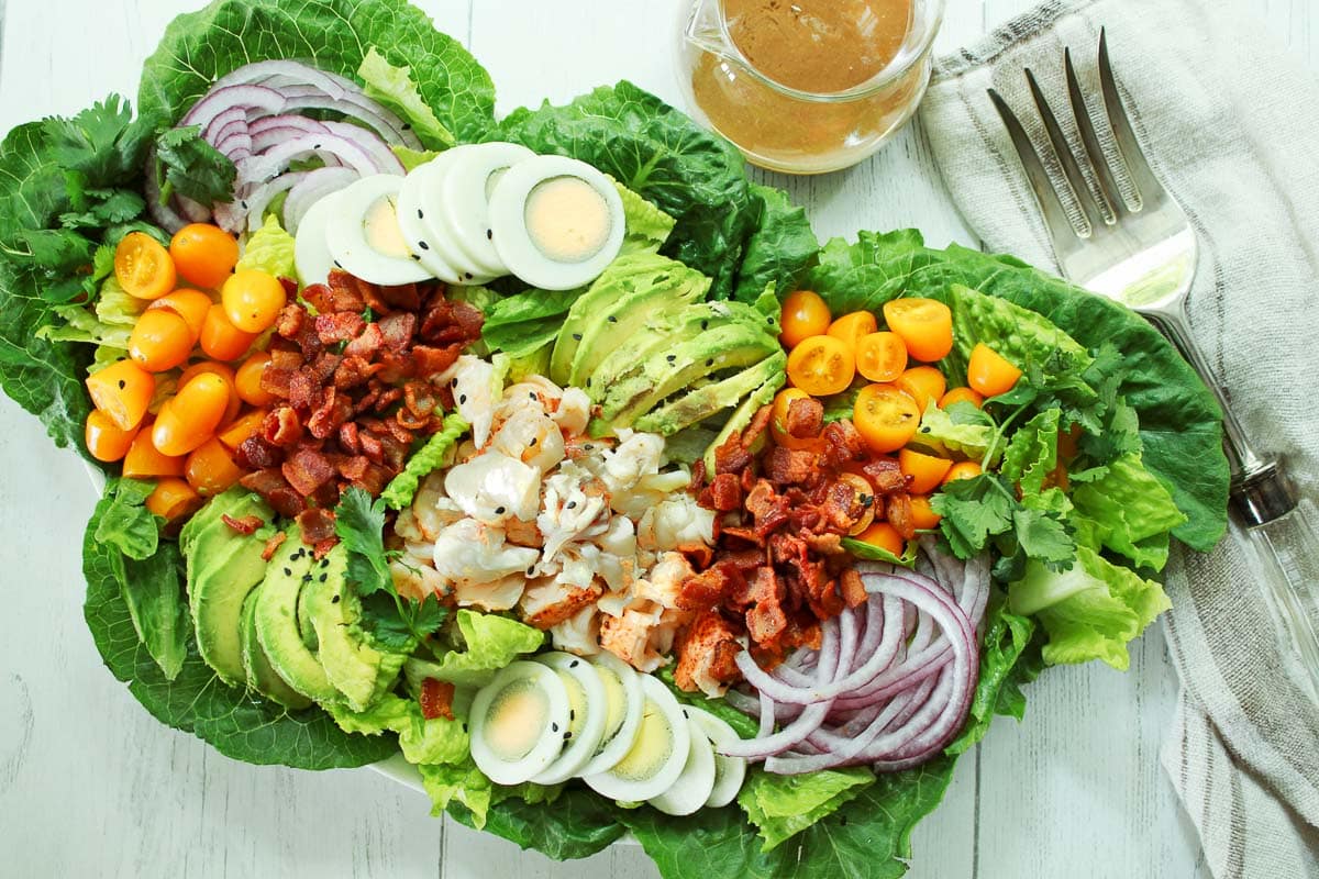 A platter of vibrant Lobster Cobb salad on top of romaine greens and a clear jar of miso dressing on the side.
